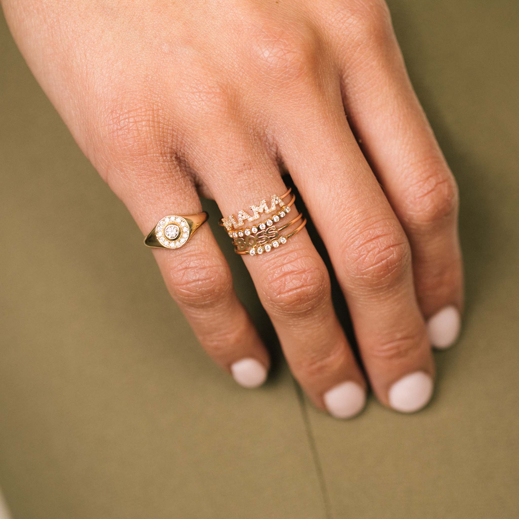 close up of woman's hand resting on her side wearing a Zoë Chicco 14kt Gold Round Diamond Halo Signet Ring on her pinky finger and an Itty Bitty Pavé Diamond MAMA Ring, Itty Bitty BOSS Ring, and a 5 and 10 Tiny Diamond Bezel Ring stacked on her ring finger