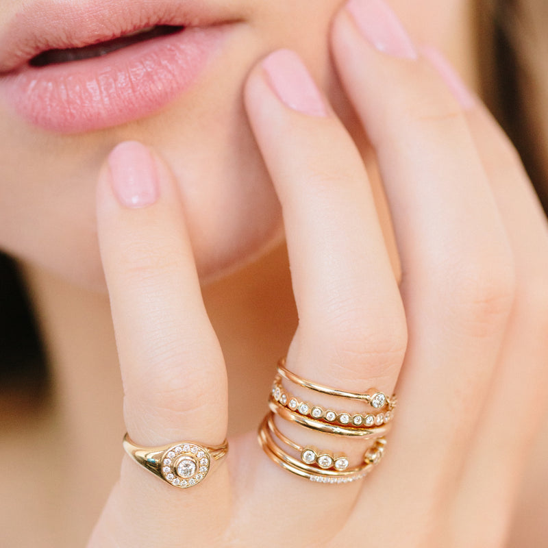 woman with hand resting on chin wearing Zoë Chicco 14kt Gold Round Diamond Halo Signet Ring on her pinky and a stack of diamond and gold rings on her fourth finger