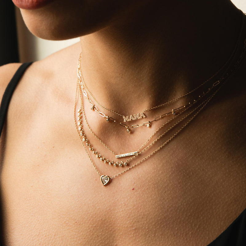 close up view of woman in a black top wearing a Zoe Chicco 14k Gold Itty Bitty Pavé Diamond MAMA Necklace layered with a 5 Dangling Diamond Bezel Small Paperclip Chain Necklace, 5 Channel Set Baguette Diamond Bar Pendant Necklace, Linked 17 Diamond Bezel Necklace, and a Floating Heart Diamond Necklace