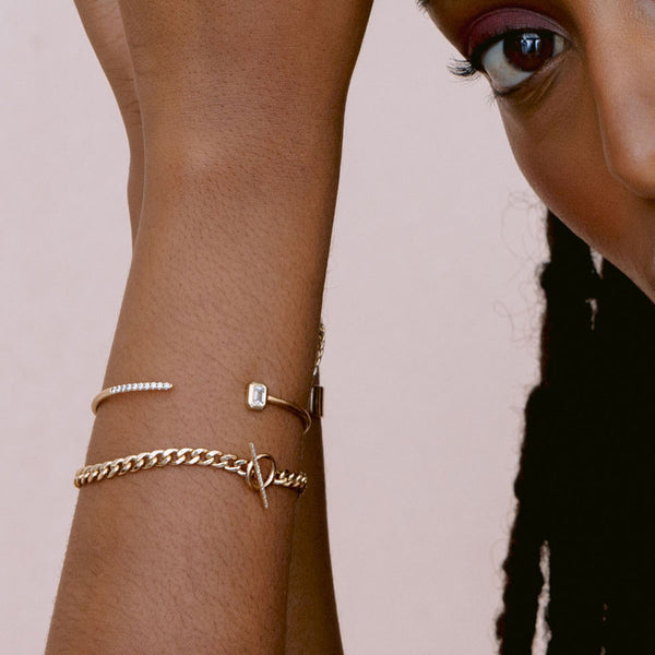 close up of woman's wrist wearing Zoë Chicco 14k Gold Medium Curb Chain Pavé Diamond Toggle Bracelet layered with a emerald cut diamond cuff bracelet