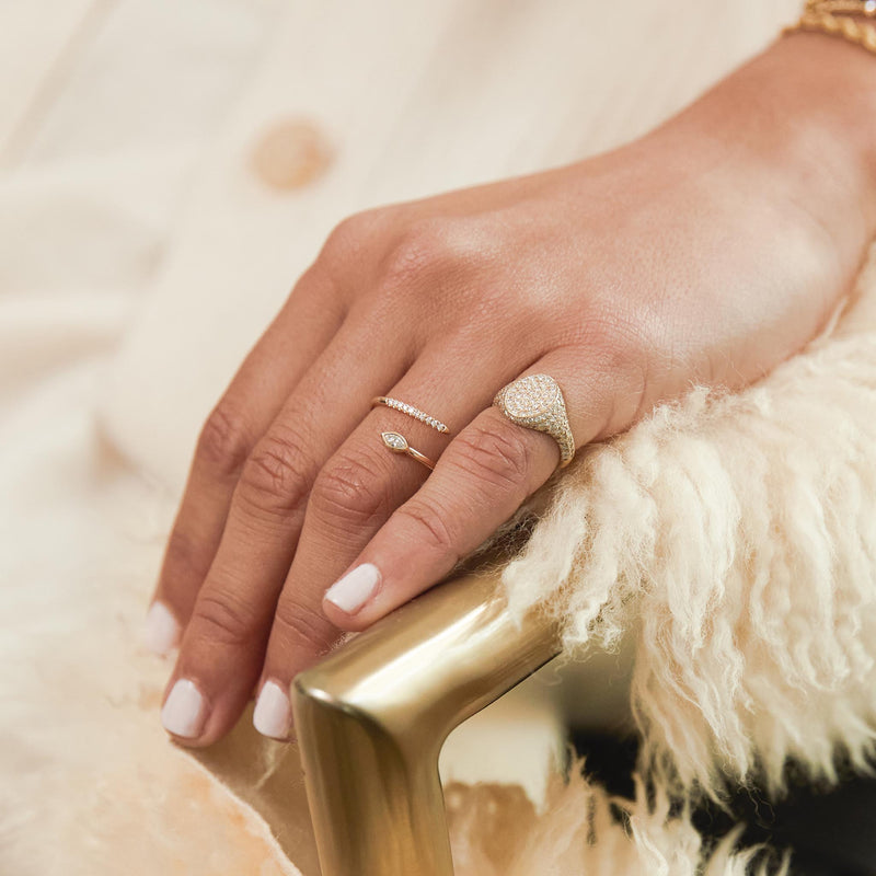 close up of a woman's hand resting on the edge of a chair wearing a Zoë Chicco 14k Gold Full Pavé Diamond Oval Signet Ring on her pinky finger and a 14k Marquise & Pavé Diamond Bypass Ring on her ring finger