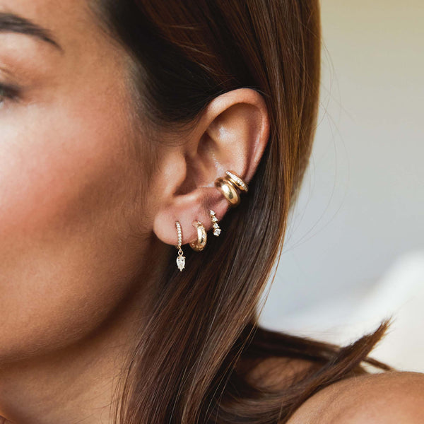 angled view of a brunette woman's ear wearing a Zoë Chicco 14k Gold Dangling Pear Diamond Small Pavé Diamond Hinge Huggie Hoop layered with two earrings and two ear cuffs
