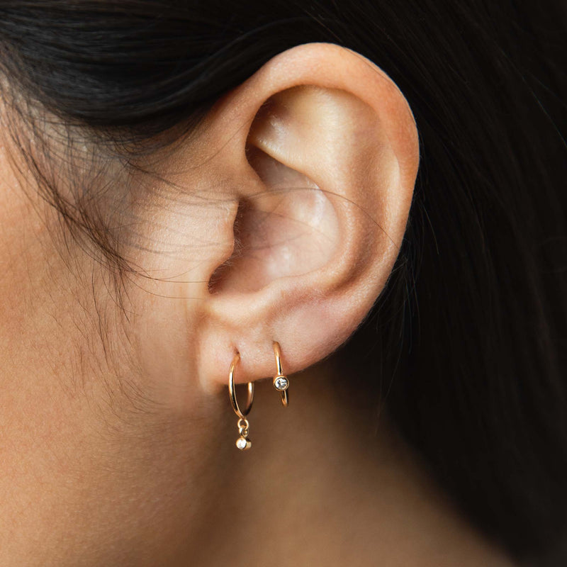 close up of woman's ear wearing a Zoë Chicco 14k Gold Diamond Bezel Thin Huggie Hoop Earring in her second piercing