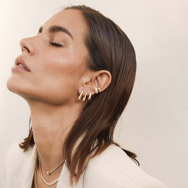 woman in a cream blazer with her eyes closed and chin raised wearing a close up of woman's ear wearing a Zoë Chicco 14k Gold Thick Large Oval Hinge Hoop Earring, Thick Medium Oval Hinge Hoop, and Pavé Diamond Thick Large Oval Hinge Hoop with two ear cuffs