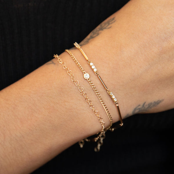 close up of a woman's wrist wearing a Zoë Chicco 14k Mixed Gold & Diamond Bar Bracelet layered with a Floating Diamond XS curb chain bracelet and a Heart Link Chain Bracelet