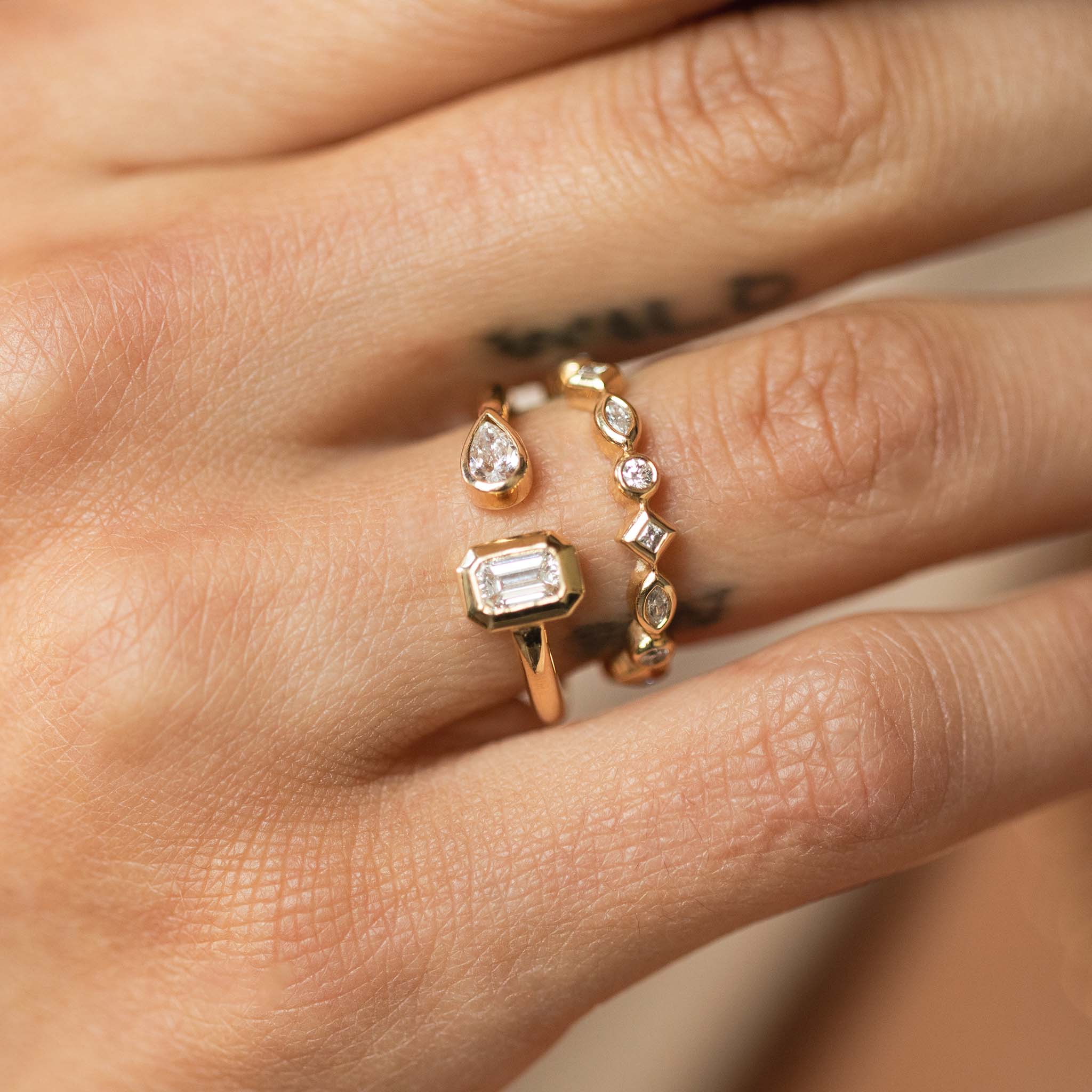 close up of a woman's hand wearing a Zoë Chicco 14k Yellow Gold Pear & Emerald Cut Diamond Bezel Open Ring stacked with a 14k Bezel Set Mixed Cut Diamond Eternity Band