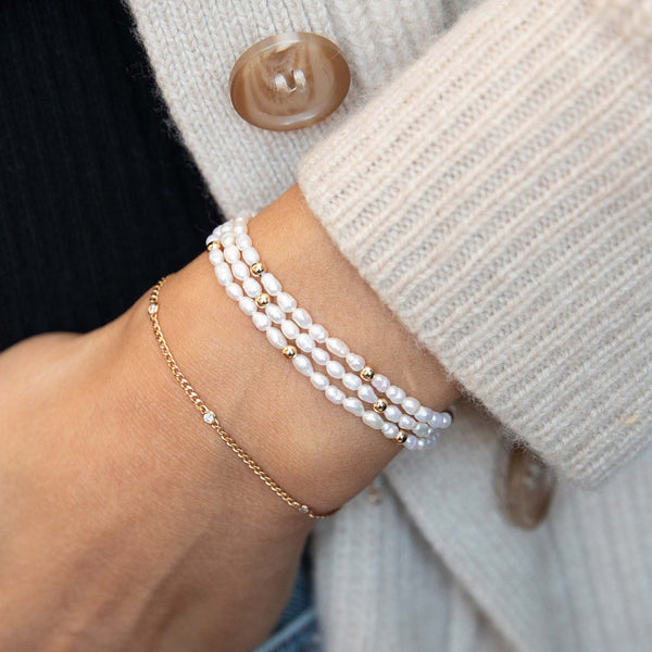 woman in a beige sweater wearing a Zoë Chicco 14k Gold XS Curb Chain Bracelet with 5 Floating Diamonds layered with a 14k Gold Bead & Rice Pearl Wrap Bracelet